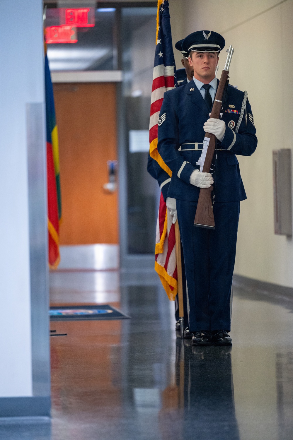 A U.S. Air Force honor guard member stands at attention in a formal indoor corridor, holding a ceremonial rifle while wearing dress blues. At least one additional honor guard member is visible behind him, and an American flag with gold fringe is positioned nearby. The setting appears to be a military or government building, likely during a formal ceremony or color guard presentation.