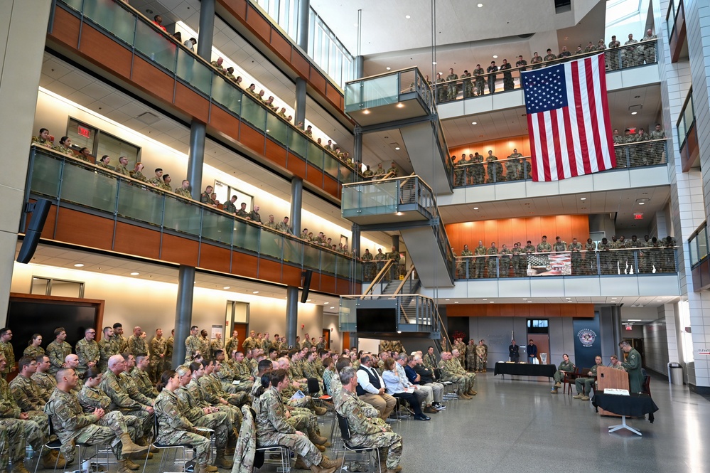 A large military formation or ceremony is taking place inside a multi-story atrium of what appears to be a military installation or government building. Hundreds of uniformed personnel in camouflage uniforms are seated on the ground floor and standing on multiple balcony levels observing a speaker at a podium. A large American flag is prominently displayed hanging from an upper balcony on the right side of the atrium.