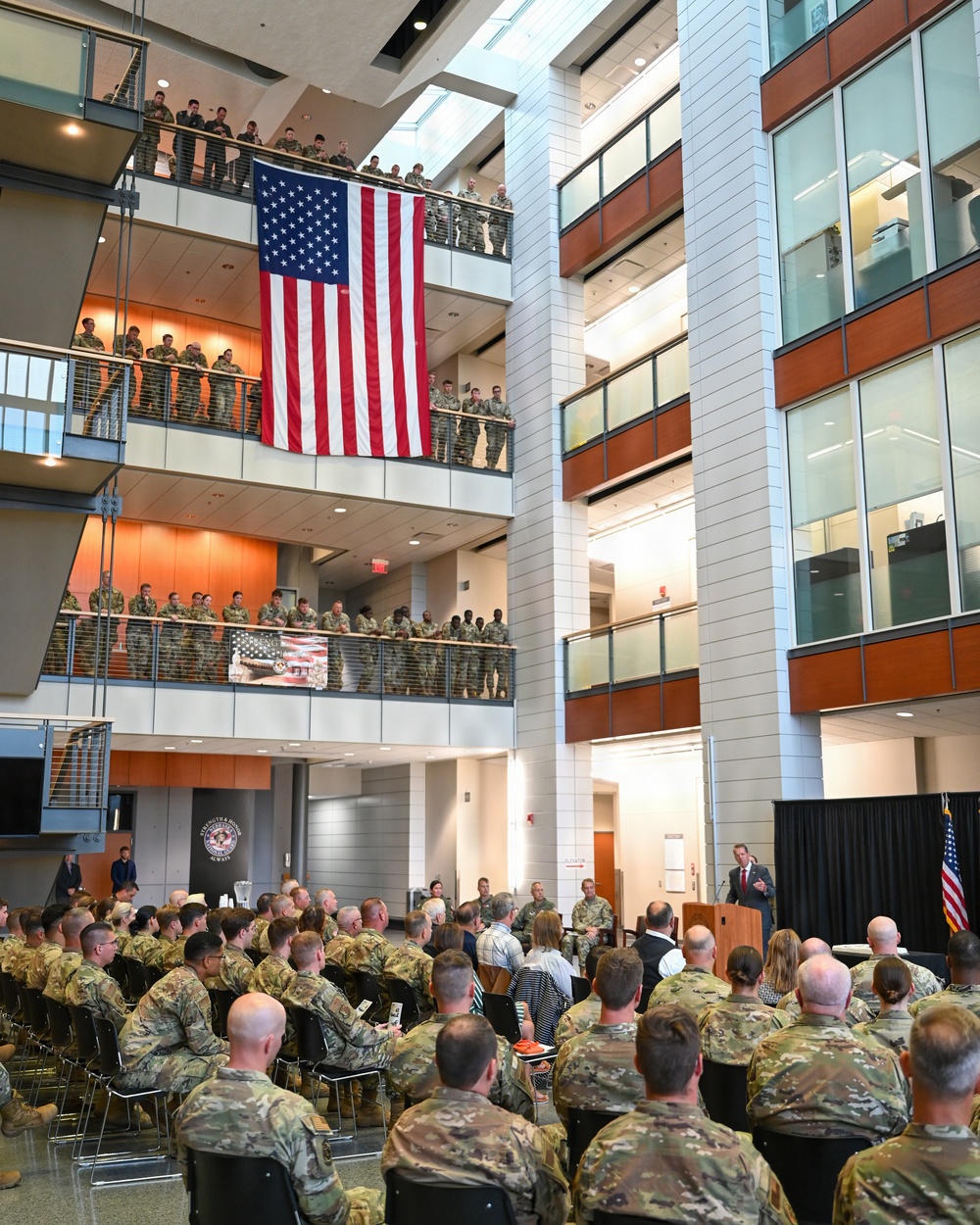 A large military ceremony or briefing is taking place inside a modern multi-story atrium building. A large American flag is prominently displayed hanging from an upper balcony, with military personnel in camouflage uniforms (OCP/MultiCam pattern) filling both the ground floor seating area and multiple upper balcony levels. A civilian speaker addresses the assembled troops from a podium near an American flag on a stand.