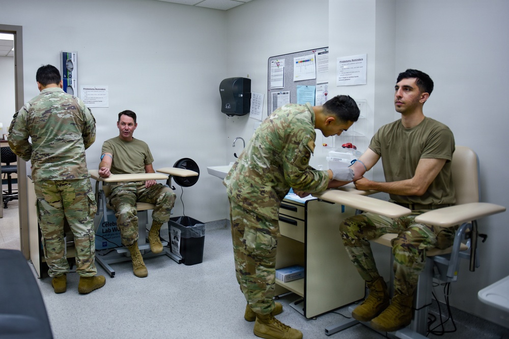 Military personnel in OCP (Operational Camouflage Pattern) uniforms are inside a medical laboratory or clinic setting. A phlebotomist in uniform is drawing blood or performing a medical procedure on a seated soldier's arm. At least two patients appear to be receiving or waiting for medical attention simultaneously.