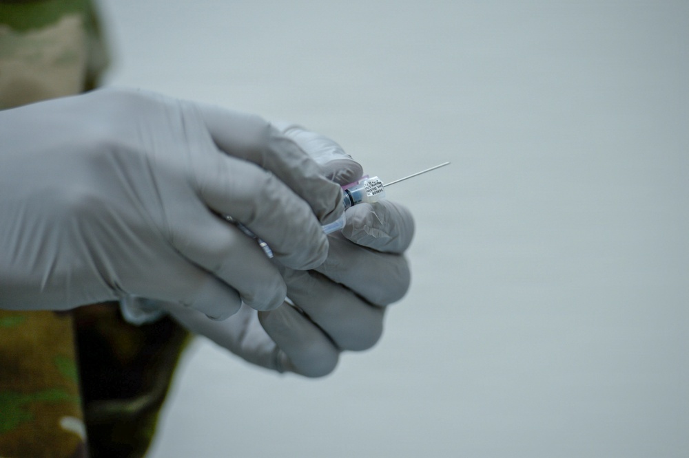 A close-up photograph of a gloved hand holding a syringe with a needle attached and a small vial. The person is wearing white latex/nitrile medical gloves and appears to be in a medical or clinical setting. A camouflage uniform sleeve is partially visible, suggesting military medical personnel.