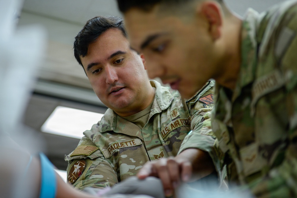 Two U.S. Air Force personnel in OCP (Operational Camouflage Pattern) uniforms are engaged in a hands-on task, with one individual named 'Paquette' appearing to instruct or work alongside the other. The image is taken from a low angle with shallow depth of field, keeping Paquette in sharp focus while the second airman is slightly blurred in the foreground. Both appear focused on a task being performed with their hands.