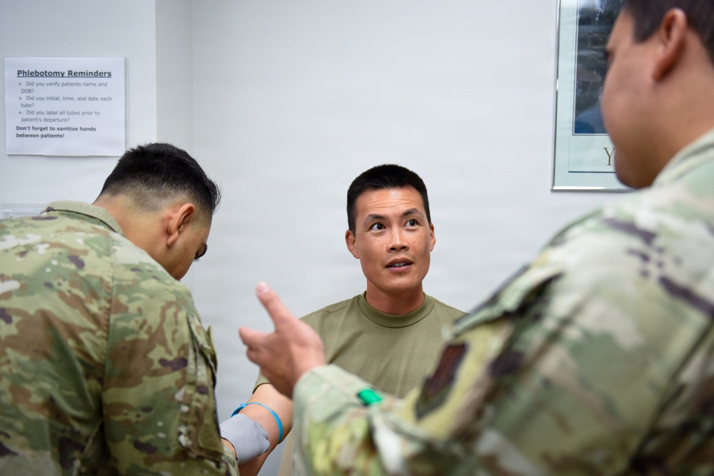Three military personnel in camouflage uniforms are gathered in what appears to be a medical phlebotomy room. The central figure, wearing an olive/tan t-shirt, appears to be a patient with a tourniquet or blood draw band on their arm, while the others appear to be medical staff. A 'Phlebotomy Reminders' checklist is posted on the wall behind them.