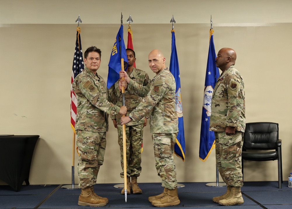 A military change of command ceremony is taking place indoors, with two officers jointly holding a unit guidon (flag on a staff) in a formal transfer gesture. Four personnel in U.S. Air Force OCP (Operational Camouflage Pattern) uniforms are present, with flags including the U.S. flag and Air Force flags displayed in the background. The setting appears to be a military installation meeting room or auditorium.