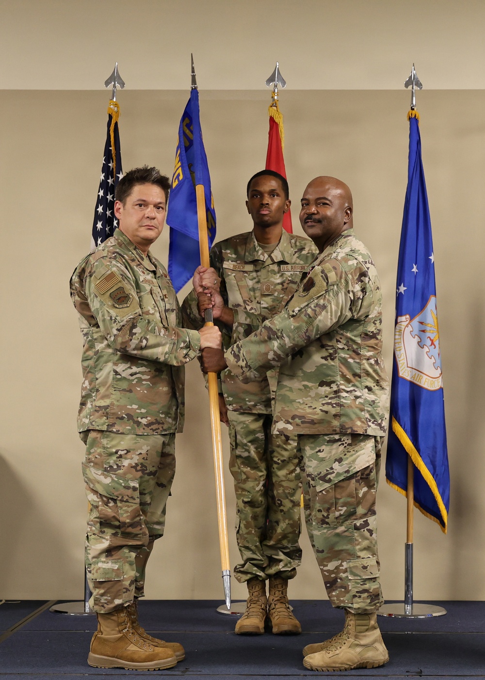 A formal U.S. Air Force change of command ceremony is taking place, with two senior personnel passing a unit guidon (flag) between them. A third service member stands in the center holding the guidon staff during the transfer. Multiple flags are displayed in the background, including the U.S. flag and the U.S. Air Force flag.