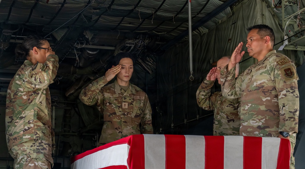 Four U.S. Air Force personnel in OCP (Operational Camouflage Pattern) uniforms are rendering a salute over a flag-draped casket inside what appears to be a military cargo aircraft. This appears to be a dignified transfer ceremony, a formal military honors procedure for returning fallen service members. The American flag is draped over the transfer case in the traditional manner.