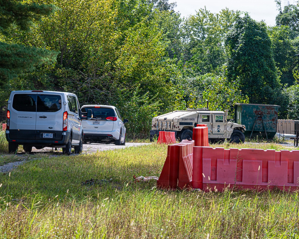 The image shows a security checkpoint or access control point on a rural road, featuring a military-style HMMWV (Humvee) marked 'Security Forces' alongside red plastic jersey barriers. Two civilian vehicles — a white Ford Transit van and a white compact car — appear to be queued or stopped at the checkpoint. The setting is surrounded by dense green trees and overgrown grass, suggesting a semi-rural or base perimeter location.