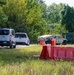 The image shows a security checkpoint or access control point on a rural road, featuring a military-style HMMWV (Humvee) marked 'Security Forces' alongside red plastic jersey barriers. Two civilian vehicles — a white Ford Transit van and a white compact car — appear to be queued or stopped at the checkpoint. The setting is surrounded by dense green trees and overgrown grass, suggesting a semi-rural or base perimeter location.