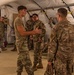 Military personnel in OCP (Operational Camouflage Pattern) uniforms are gathered inside a large military tent or temporary structure. A soldier in a tan t-shirt and OCP trousers appears to be briefing or explaining something to fellow service members. The setting suggests a field operations environment or training exercise.