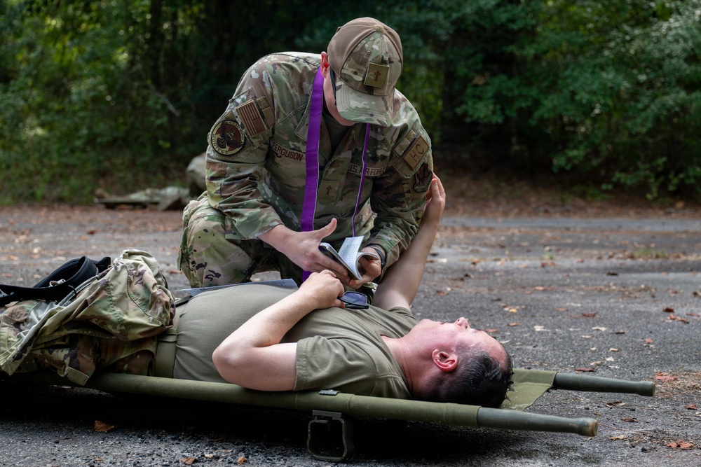 A military service member in OCP (Operational Camouflage Pattern) uniform, identified by name tape as 'Ferguson,' is performing a medical assessment or treatment on a casualty lying on a litter/stretcher on a paved surface. The standing individual is wearing a purple sash/ribbon, which typically designates a role-player or evaluator in a training exercise. The scene appears to be a tactical combat casualty care (TCCC) training scenario conducted outdoors near a wooded area.