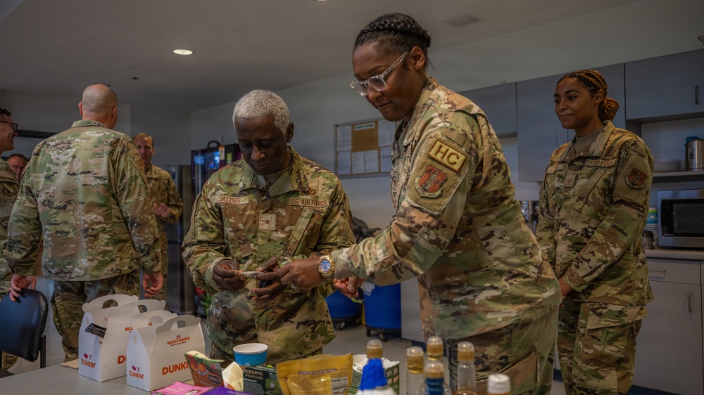 A group of U.S. Air Force personnel in OCP (Operational Camouflage Pattern) uniforms are gathered in what appears to be a break room or common area. Two central figures, one identified by nametape as 'JACOBS' and another wearing an 'HC' (Chaplain) tab, are examining items on a table stocked with snacks and beverages. Several other uniformed personnel are visible in the background.