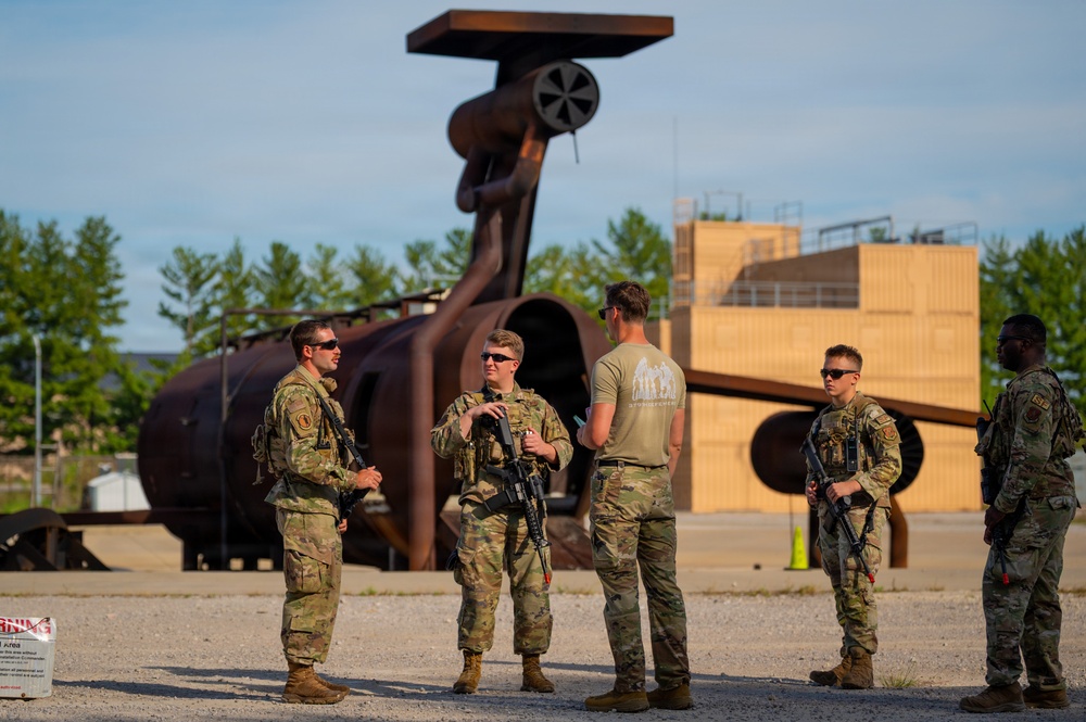 Five military personnel in camouflage uniforms are gathered in an outdoor training or industrial area, appearing to receive a briefing or conduct a debrief. Four of the individuals are armed with rifles and wearing full combat gear, while one person in a tan t-shirt appears to be an instructor or evaluator. A large rusted industrial or training structure is visible in the background along with a wooden building.