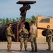 Five military personnel in camouflage uniforms are gathered in an outdoor training or industrial area, appearing to receive a briefing or conduct a debrief. Four of the individuals are armed with rifles and wearing full combat gear, while one person in a tan t-shirt appears to be an instructor or evaluator. A large rusted industrial or training structure is visible in the background along with a wooden building.
