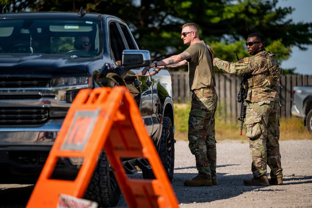 Two military personnel in MultiCam/OCP camouflage uniforms are conducting a vehicle checkpoint inspection on a gravel road. One soldier is handing or receiving documents from the driver of a dark-colored Chevrolet pickup truck, while the second soldier stands in a security overwatch position carrying a rifle. An orange safety marker/sign is visible in the foreground.