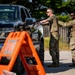 Two military personnel in MultiCam/OCP camouflage uniforms are conducting a vehicle checkpoint inspection on a gravel road. One soldier is handing or receiving documents from the driver of a dark-colored Chevrolet pickup truck, while the second soldier stands in a security overwatch position carrying a rifle. An orange safety marker/sign is visible in the foreground.