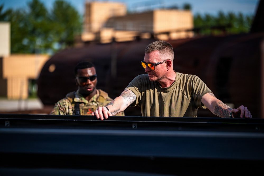 Two individuals are working together near what appears to be a military or industrial vehicle, with one person in the foreground wearing an olive green t-shirt and orange-tinted sunglasses with visible tattoos on his arms. The second person in the background is wearing military camouflage (OCP/MultiCam pattern) uniform and dark sunglasses. The setting appears to be an outdoor industrial or military training area during golden hour lighting.