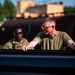 Two individuals are working together near what appears to be a military or industrial vehicle, with one person in the foreground wearing an olive green t-shirt and orange-tinted sunglasses with visible tattoos on his arms. The second person in the background is wearing military camouflage (OCP/MultiCam pattern) uniform and dark sunglasses. The setting appears to be an outdoor industrial or military training area during golden hour lighting.