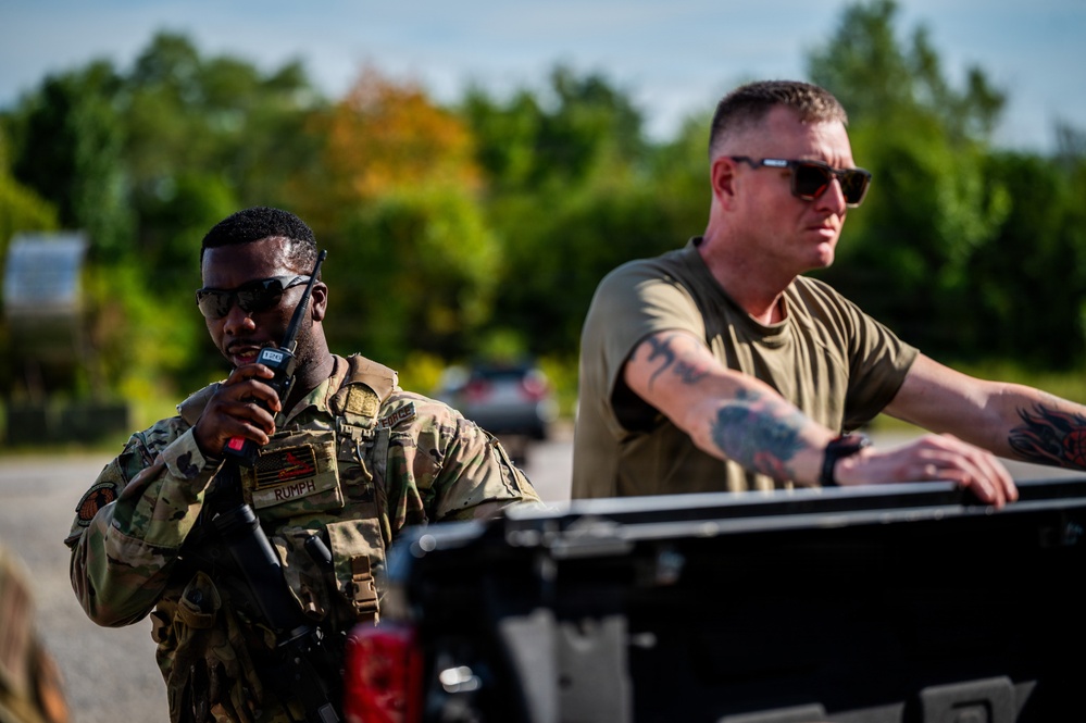 Two military personnel are visible in an outdoor setting near a vehicle. The individual on the left is in full camouflage uniform with a name tape reading 'RUMPH' and is speaking into a radio while carrying a weapon. The individual on the right is in a military olive green t-shirt with visible tattoos on his arm, leaning against what appears to be a pickup truck bed.