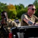 Two military personnel are visible in an outdoor setting near a vehicle. The individual on the left is in full camouflage uniform with a name tape reading 'RUMPH' and is speaking into a radio while carrying a weapon. The individual on the right is in a military olive green t-shirt with visible tattoos on his arm, leaning against what appears to be a pickup truck bed.