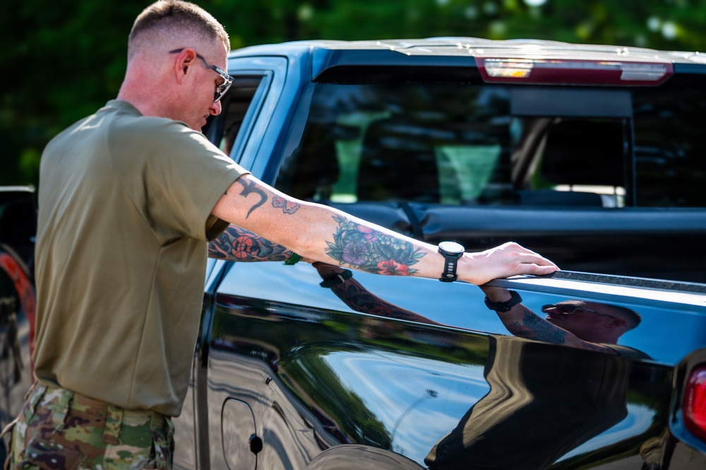 A military service member in olive drab t-shirt and camouflage pants is leaning over the bed of a black pickup truck. The individual has extensive tattoos on their right forearm and is wearing sunglasses and a watch. The setting appears to be an outdoor area with green trees visible in the background.