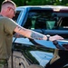 A military service member in olive drab t-shirt and camouflage pants is leaning over the bed of a black pickup truck. The individual has extensive tattoos on their right forearm and is wearing sunglasses and a watch. The setting appears to be an outdoor area with green trees visible in the background.