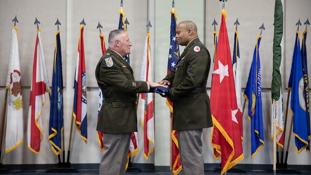 Two U.S. Army officers in green service uniforms are participating in a formal military ceremony, exchanging a folded American flag. The setting features a backdrop of multiple state and military flags on staffs with gold fringe and spear-tip finials. This appears to be a change of command, retirement, or similar official military ceremony.