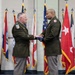 Two U.S. Army officers in green service uniforms are participating in a formal military ceremony, exchanging a folded American flag. The setting features a backdrop of multiple state and military flags on staffs with gold fringe and spear-tip finials. This appears to be a change of command, retirement, or similar official military ceremony.