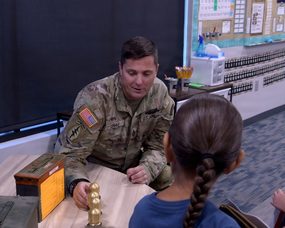 A U.S. Army soldier in MultiCam/OCP uniform is seated at a table in what appears to be an elementary school classroom, engaging with a young girl. The soldier, whose uniform displays 'U.S. ARMY,' an Airborne tab, and an American flag patch, appears to be showing the child a brass-colored object, possibly an inert training round or display item. The setting suggests a school visit or career day presentation.