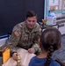 A U.S. Army soldier in MultiCam/OCP uniform is seated at a table in what appears to be an elementary school classroom, engaging with a young girl. The soldier, whose uniform displays 'U.S. ARMY,' an Airborne tab, and an American flag patch, appears to be showing the child a brass-colored object, possibly an inert training round or display item. The setting suggests a school visit or career day presentation.