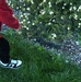 A person wearing a red hoodie, black pants, and black-and-white Nike sneakers is standing on a green lawn near a garden border. The garden features a flowering shrub with small white blooms, river rocks, and a black plastic drainage grate. The photo is taken from an overhead angle, showing only the lower body of the individual.