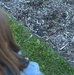 A person with long reddish-brown hair is looking down at a ground-level scene consisting of wood chip mulch and a grass border. A dark-colored caterpillar or larva is visible among the wood chips near the grass edge. The perspective is from above, shot over the person's shoulder toward the ground.