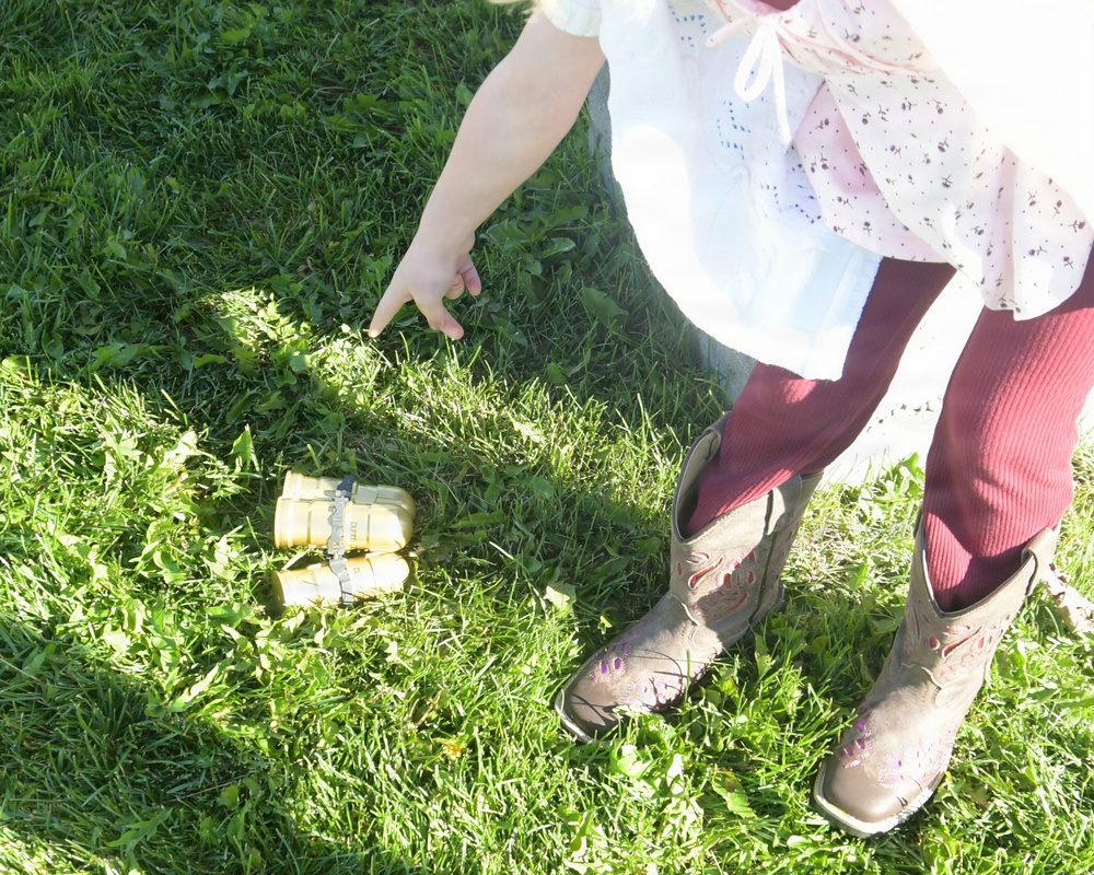 A young child wearing a white dress with floral print, dark red/burgundy leggings, and brown embroidered cowboy boots is standing on green grass. The child is pointing downward toward a small object on the ground, which appears to be a tiny pair of toy or miniature boots. The scene is captured from an overhead angle in bright natural sunlight.