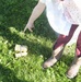 A young child wearing a white dress with floral print, dark red/burgundy leggings, and brown embroidered cowboy boots is standing on green grass. The child is pointing downward toward a small object on the ground, which appears to be a tiny pair of toy or miniature boots. The scene is captured from an overhead angle in bright natural sunlight.