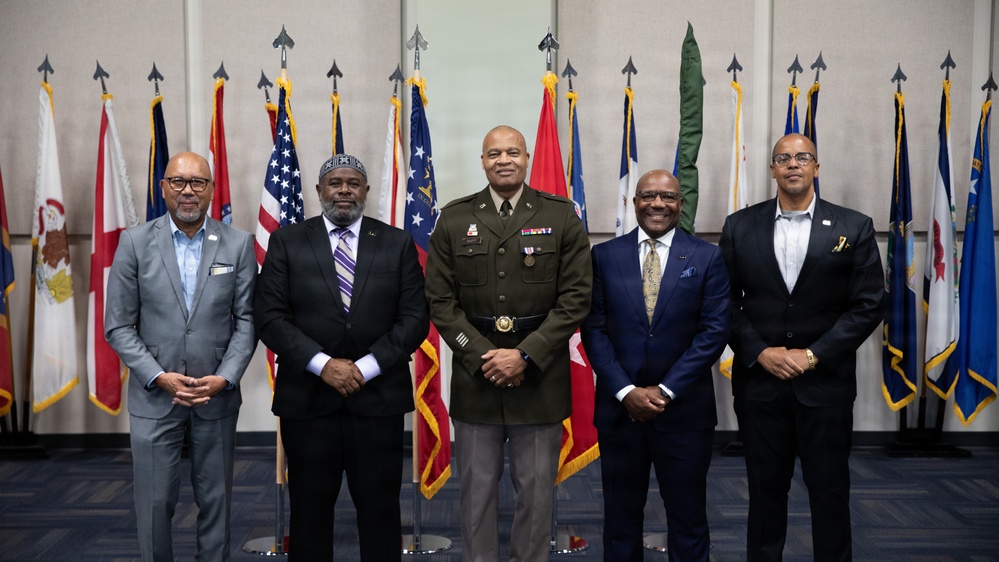 Five men pose for a formal group photograph in front of a display of multiple flags, including the United States flag and various state and military flags. The central figure is wearing a U.S. Army dress uniform with medals and rank insignia, while the other four men are dressed in civilian business attire. The setting appears to be an official military or government facility.