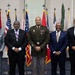 Five men pose for a formal group photograph in front of a display of multiple flags, including the United States flag and various state and military flags. The central figure is wearing a U.S. Army dress uniform with medals and rank insignia, while the other four men are dressed in civilian business attire. The setting appears to be an official military or government facility.