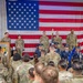 A formal military ceremony is taking place indoors, likely a change of command or unit activation event. Personnel in OCP (Operational Camouflage Pattern) uniforms stand at attention on a stage backdrop by a large American flag, while an Air Force Honor Guard presents colors to an assembled formation. The setting includes the Arizona state flag and a POW/MIA flag, suggesting a National Guard or Reserve installation in Arizona.