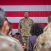 A U.S. Army soldier stands at attention in front of a large American flag backdrop, appearing to address or be observed by a group of personnel. The central figure is wearing OCP (Operational Camouflage Pattern) uniform with visible rank insignia and name tape. The scene appears to be a formal military ceremony or formation event.