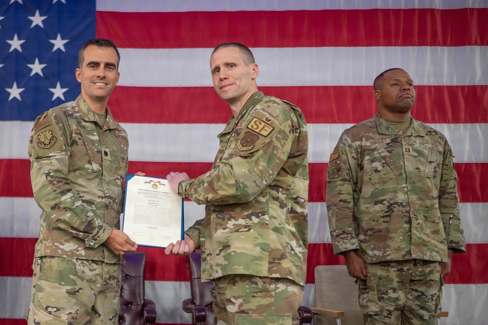 Three military personnel in OCP (Operational Camouflage Pattern) uniforms are posed in front of a large American flag during what appears to be a formal awards or recognition ceremony. Two individuals in the foreground are jointly holding a framed certificate or citation document, suggesting a medal or commendation presentation. A third service member stands at attention in the background to the right.