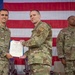 Three military personnel in OCP (Operational Camouflage Pattern) uniforms are posed in front of a large American flag during what appears to be a formal awards or recognition ceremony. Two individuals in the foreground are jointly holding a framed certificate or citation document, suggesting a medal or commendation presentation. A third service member stands at attention in the background to the right.