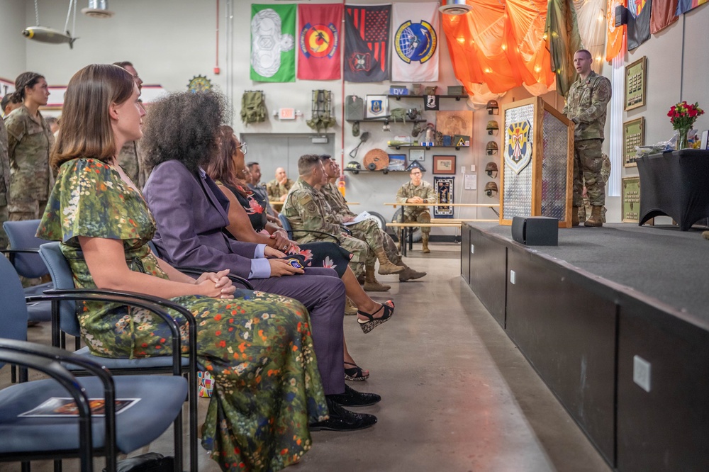 A military ceremony or briefing is taking place in what appears to be a unit heritage room or common area on a military installation. An airman in camouflage uniform stands at a podium addressing an audience of both military personnel in uniform and civilian guests seated in rows. The room is decorated with various military unit flags, patches, helmets, and memorabilia displayed on the walls.