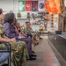 A military ceremony or briefing is taking place in what appears to be a unit heritage room or common area on a military installation. An airman in camouflage uniform stands at a podium addressing an audience of both military personnel in uniform and civilian guests seated in rows. The room is decorated with various military unit flags, patches, helmets, and memorabilia displayed on the walls.