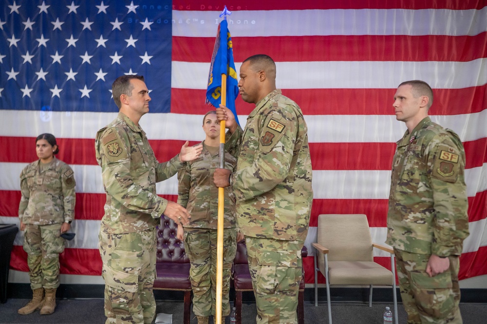 A military change of command ceremony is taking place in front of a large American flag backdrop. A guidon (unit flag) is being passed between two service members, which is the central symbolic act of a change of command. The personnel are wearing U.S. Air Force OCP (Operational Camouflage Pattern) uniforms with 'SF' (Security Forces) patches visible.