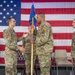 A military change of command ceremony is taking place in front of a large American flag backdrop. A guidon (unit flag) is being passed between two service members, which is the central symbolic act of a change of command. The personnel are wearing U.S. Air Force OCP (Operational Camouflage Pattern) uniforms with 'SF' (Security Forces) patches visible.