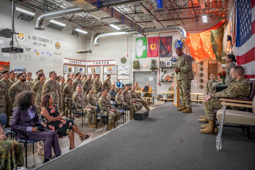 A military ceremony or briefing is taking place inside a facility identified as 'The Foundry' with a 'Combat Corner' display area visible on the wall. A senior military officer in Army Combat Uniform (ACU) stands at a podium addressing a large formation of soldiers, while two civilian women in formal attire are seated in the foreground as guests of honor. Multiple military unit flags and an American flag are displayed prominently in the background.