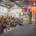 A military ceremony or briefing is taking place inside a facility identified as 'The Foundry' with a 'Combat Corner' display area visible on the wall. A senior military officer in Army Combat Uniform (ACU) stands at a podium addressing a large formation of soldiers, while two civilian women in formal attire are seated in the foreground as guests of honor. Multiple military unit flags and an American flag are displayed prominently in the background.