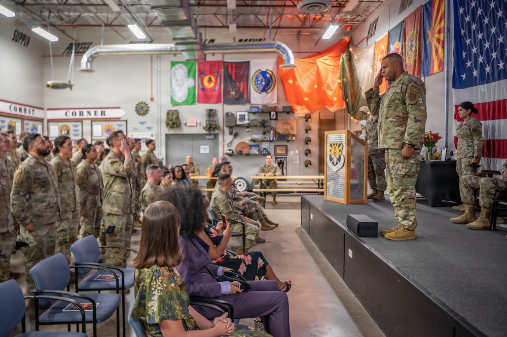 A military ceremony is taking place inside what appears to be a National Guard or military unit facility, likely a promotion, retirement, or change of command ceremony. Uniformed service members are rendering a salute toward a senior officer standing on a raised stage near a podium, while civilian family members are seated in the foreground. Multiple military unit flags and the U.S. flag are displayed on the walls, along with a 'Combat Corner' display.