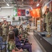 A military ceremony is taking place inside what appears to be a National Guard or military unit facility, likely a promotion, retirement, or change of command ceremony. Uniformed service members are rendering a salute toward a senior officer standing on a raised stage near a podium, while civilian family members are seated in the foreground. Multiple military unit flags and the U.S. flag are displayed on the walls, along with a 'Combat Corner' display.