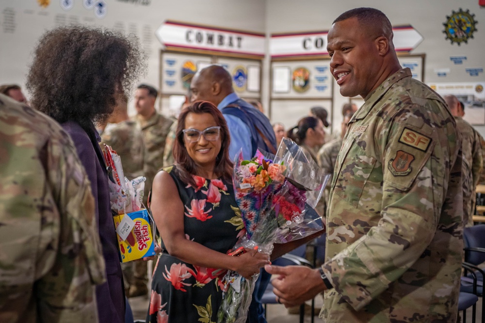 A military ceremony or celebration is taking place in what appears to be a military facility with 'COMBAT CO' signage visible in the background. A smiling airman in OCP (Operational Camouflage Pattern) uniform with an 'SF' (Security Forces) patch is interacting with civilians, one of whom is holding flowers and a box of Swedish Fish candy. The atmosphere appears festive and celebratory, likely a homecoming, promotion, or recognition event.