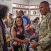 A military ceremony or celebration is taking place in what appears to be a military facility with 'COMBAT CO' signage visible in the background. A smiling airman in OCP (Operational Camouflage Pattern) uniform with an 'SF' (Security Forces) patch is interacting with civilians, one of whom is holding flowers and a box of Swedish Fish candy. The atmosphere appears festive and celebratory, likely a homecoming, promotion, or recognition event.