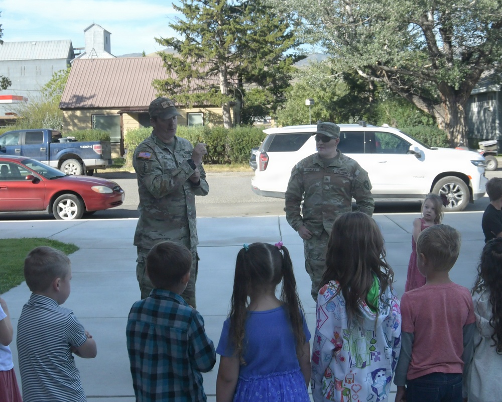 Two U.S. Army soldiers in camouflage uniforms are speaking to a group of young children outdoors on a paved surface. The setting appears to be a community or school event where military personnel are engaging with children, possibly for an educational or outreach visit. The scene takes place in a small town or suburban area with buildings, trees, and parked vehicles visible in the background.